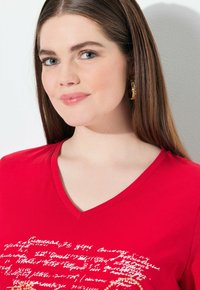 Camiseta roja de cuello en V con diseño de texto en blanco y dorado. Tejido suave con mangas cortas, estilo minimalista y escote simple.