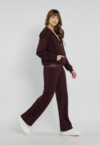 Burgundowy hoodie z troczkiem i przednimi kieszeniami, zestawiony z pasującymi spodniami o szerokich nogawkach. Białe trampki dopełniają outfit na jasnym tle.