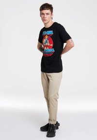 Camiseta de algodón negra con un gráfico colorido de He-Man. Combinada con pantalones claros tapered beige y zapatillas negras.