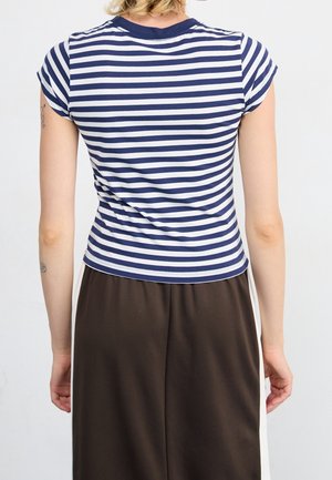 Personne portant une chemise à manches courtes rayée bleu marine et blanc et un pantalon brun foncé, vue de dos sur un fond blanc.