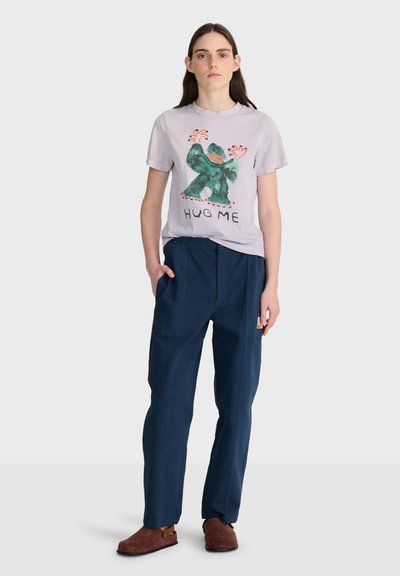 Femme debout, main dans la poche, portant un T-shirt violet clair avec une créature verte et le texte "HUG ME", pantalon bleu marine et chaussons bruns.