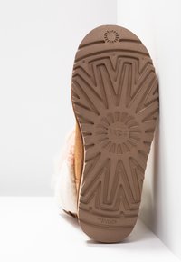Podeszwa butów UGG wykonana z teksturowanego materiału gumowego. Zawiera wzór w zygzaki oraz okrągłe logo na środku. Kolor: jasnobrązowy.