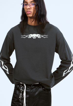 Chemise à manches longues gris foncé avec un graphique de skull blanc et des motifs de flammes sur les manches ; assortie avec un pantalon en cuir noir et des accessoires.