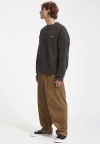 Ciemnozielona bluza z okrągłym dekoltem i białym logo na piersi, w połączeniu z luźnymi brązowymi spodniami z kordurowej tkaniny i czarnymi sneakersami.