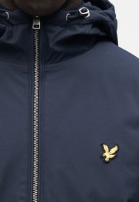 Mørkeblå jakke med en sølvfarvet lynlås foran, en hætte med snoretræk og en lille gul fuglelogo på brystet.