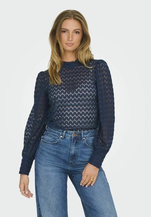 Blouse transparente bleu marine avec un motif en zigzag, manches longues bouffantes et encolure ronde, associée à un jean en denim taille haute.
