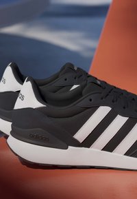 Scarpe da ginnastica Adidas bianche e nere con tre strisce bianche, lacci neri e suole intermedie bianche, posizionate su una superficie arancione all'aperto.