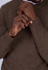 Chandail en tricot marron avec des poignets côtelés, mettant en avant une texture douce. Des mains ajustent le col, soulignant le design confortable et décontracté du vêtement.