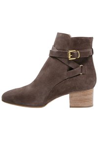 Bottine en daim marron à bout pointu, avec des sangles décoratives à boucle et un talon empilé. Texture lisse et design minimaliste.