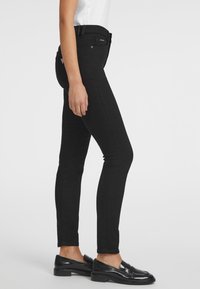 Crne uske traperice visokog struka, slim fit, s klasičnim zatvaranjem na dugme, prikazane na bočnom profilu modela.