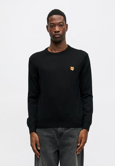 Ung mann iført en svart genser med et brodert oransje revhode på venstre bryst, kombinert med falmede svarte jeans.