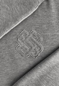 Grijze stof met een gestikt geometrisch patroon in het midden, met in elkaar grijpende vormen en zichtbare naden.