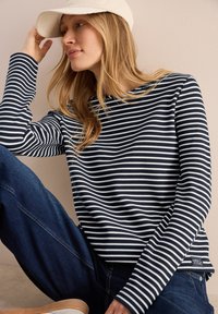 Langärmliges Oberteil mit navy-blauen und weißen Streifen sowie einem Bootsausschnitt, kombiniert mit dunkelblauen Jeans und einer hellen Mütze.