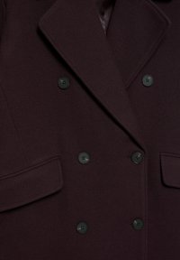 Donkerpaarse double-breasted wollen jas met zes zwarte knopen en twee klepzakken, voorzien van een brede reverskraag met inkeping.