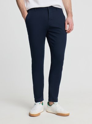 Mężczyzna noszący dopasowane granatowe spodnie i białe trampki z zielonymi akcentami, stojący z rękami w kieszeniach.