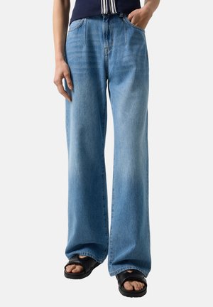 Persoon die lichtblauwe jeans met wijde pijpen en zwarte sandalen met open teen draagt, staand met één hand in de zak en de andere ontspannen langs het lichaam.