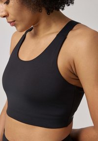 Reggiseno sportivo nero con una texture liscia, caratterizzato da spalline larghe, scollo rotondo e design corto. Stile minimalista e funzionale.