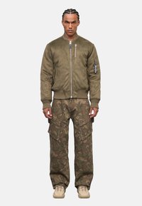 Olivgrüne Bomberjacke mit Reißverschluss und strukturiertem Stoff; kombiniert mit Camouflage-Cargohosen mit großen Taschen und lockerer Passform.