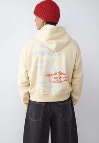 Sudadera corta color crema con capucha ajustable con cordón y gráficos de texto azul en la espalda, combinada con pantalones anchos de mezclilla oscura. Gorro de punto rojo.