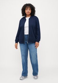 Navy zip-up cardigan with ribbed cuffs, paired with a white t-shirt and blue jeans. Features a relaxed fit and soft texture.