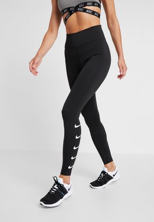 Leggings noirs en matière extensible avec taille haute, ornés de logos Nike blancs le long des jambes. Associés à des chaussures de sport noires.