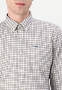 Chemise à carreaux de couleur claire avec un col boutonné, arborant des carreaux bleus, bruns et blancs. Petit logo sur la poche poitrine gauche.