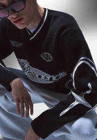 Svart stickad tröja med vita detaljer, med v-ringning, logobrodyr och grafiska mönster på ärmarna. Bärs med gråa sweatpants.