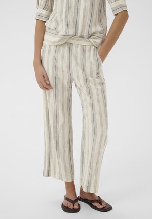Žena nosi krémové voľné pruhované nohavice a zladený top s tromištvrtinovými rukávmi, spojené s tmavohnedými žabkami.