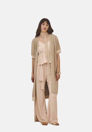 Larga cardigan abierta con un patrón a rayas beige sobre una blusa beige clara y pantalones de pierna ancha. Incluye una bufanda y pulsera a juego.
