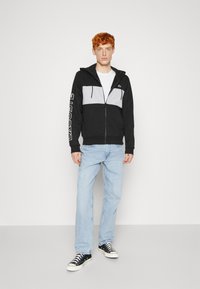 Black and gray zip-up hoodie with a front pocket, white drawstrings, and logo detail on the sleeve, worn with light blue jeans and black sneakers.