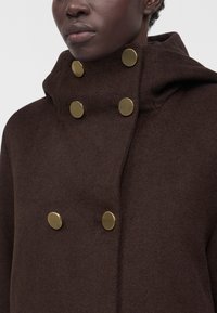 Femme portant un manteau marron foncé à col montant avec deux rangées de grands boutons ronds dorés.