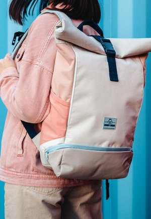 Person iført en beige og ferskenfarvet rektangulær rygsæk med sort spænde og lynlås, iført en pink jakke og beige bukser.