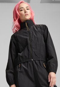 Veste noire légère, cropped, avec un col montant, fermeture éclair à l'avant, taille élastique et accents dorés sur les poches. Tissu texturé.