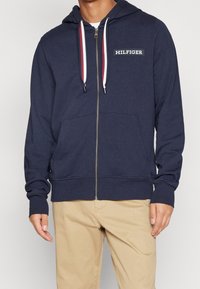 Felpa con zip di colore navy realizzata in tessuto morbido, dotata di tasca frontale e cappuccio con cordino. Presenta dettagli a contrasto in rosso e bianco.