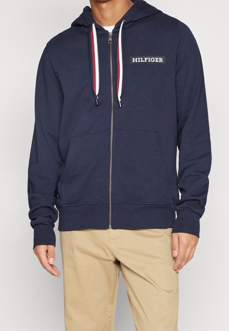 Felpa con zip di colore navy realizzata in tessuto morbido, dotata di tasca frontale e cappuccio con cordino. Presenta dettagli a contrasto in rosso e bianco.