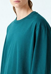 Femme portant un t-shirt à manches longues et col rond de couleur sarcelle, montrée du cou jusqu'au milieu du torse sur un fond clair uni.