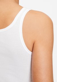 White cotton tank top with a smooth texture, featuring narrow shoulder straps and a scoop neckline, showing a glimpse of shoulder skin.