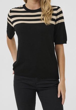 Femme portant un pull noir à manches courtes avec des rayures horizontales beiges sur le haut de la poitrine et les épaules, associé à un pantalon noir.