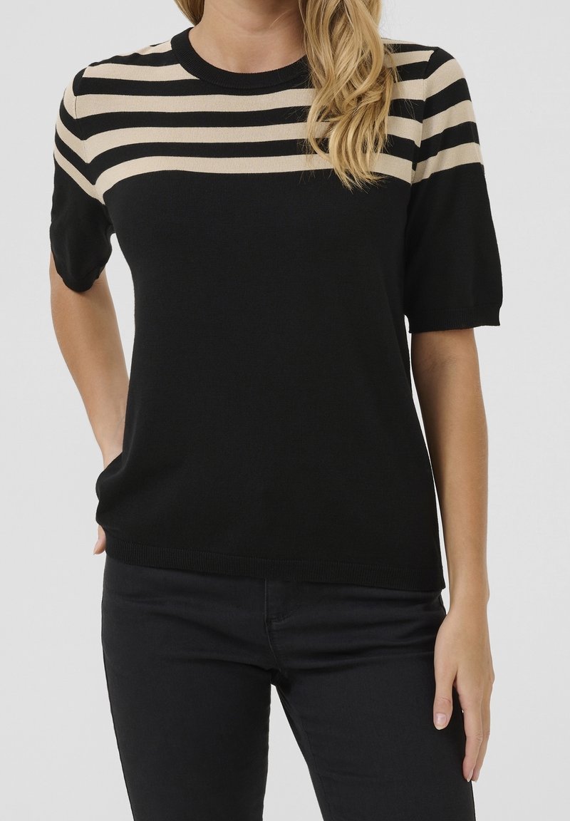 Femme portant un pull noir à manches courtes avec des rayures horizontales beiges sur le haut de la poitrine et les épaules, associé à un pantalon noir.