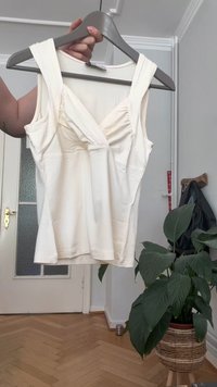 White fitted tank top with wide straps, pleated front design, smooth texture, and a straight hem, displayed on a gray hanger.