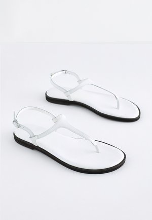 Vita platta sandaler med T-rem design, tillverkade av slätt material, med en smal ankelrem och en svart sula. Minimalistisk stil.