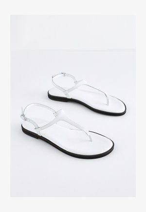 Sandales plates blanches avec un design à lanière T, fabriquées en matériau lisse, dotées d'une fine bride de cheville et d'une semelle noire. Style minimaliste.