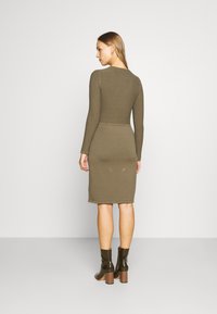 Vestido de malha canelada em verde oliva, com mangas longas, silhueta ajustada e comprimento até ao joelho. Combinado com botas pretas brilhantes até ao tornozelo com saltos quadrados.