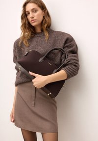 Barna kézitáska sima felülettel, dupla füllel és arany színű fémszerelvényekkel. A modellen kötött barna pulóver és sötét szoknya van.