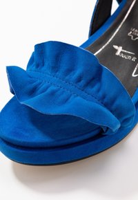 Sandale en suedine bleue avec une bride froncée, semelle rembourrée, forme incurvée et semelle noire, conçue pour le confort.