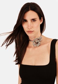 Frau mit langen dunklen Haaren, die ein schwarzes ärmelloses Oberteil und eine große silberne geknotete Choker-Halskette trägt, nach vorn gerichtet mit neutralem Ausdruck.