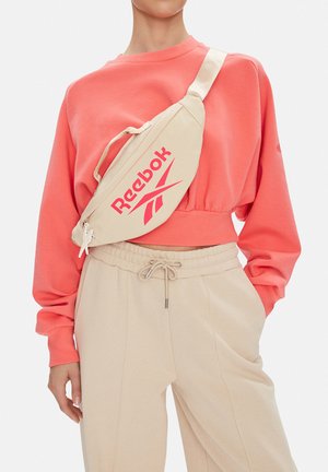 Osoba ubrana w koralową krótką bluzę i beżowe spodnie dresowe, z beżową torbą Reebok noszoną na skos przez klatkę piersiową.