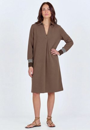 Žena stojaca na priamom pozadí, oblečená v hnedých dlhých rukávoch, dĺžka šiat po kolená so pásikovanými manžetami a béžovými plochými sandálmi na remienky.