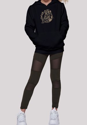 Sort hættetrøje med et guld Avengers-logo, parret med olivengrønne leggins med gennemsigtige paneler. Modellen har hvide træningssko på.