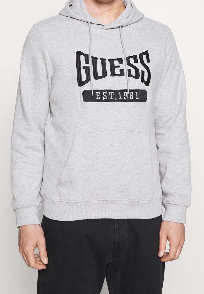 Sudadera gris con capucha, presenta un logo negro de "GUESS" y el texto "EST.1981". Incluye un bolsillo tipo canguro en la parte frontal y puños acanalados.